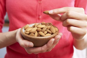 Woman in a red cardigan holding up and eating from a wooden bowl of almonds Woman in a red cardigan holding up and eating from a wooden bowl of almonds