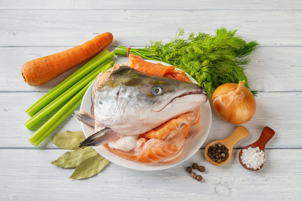 Raw ingredients for cooking fish stock: fresh salmon head and bones, vegetables, salt and pepper over a white wood table. Red fish broth bouillon recipe. Homemade seafood healthy eating.