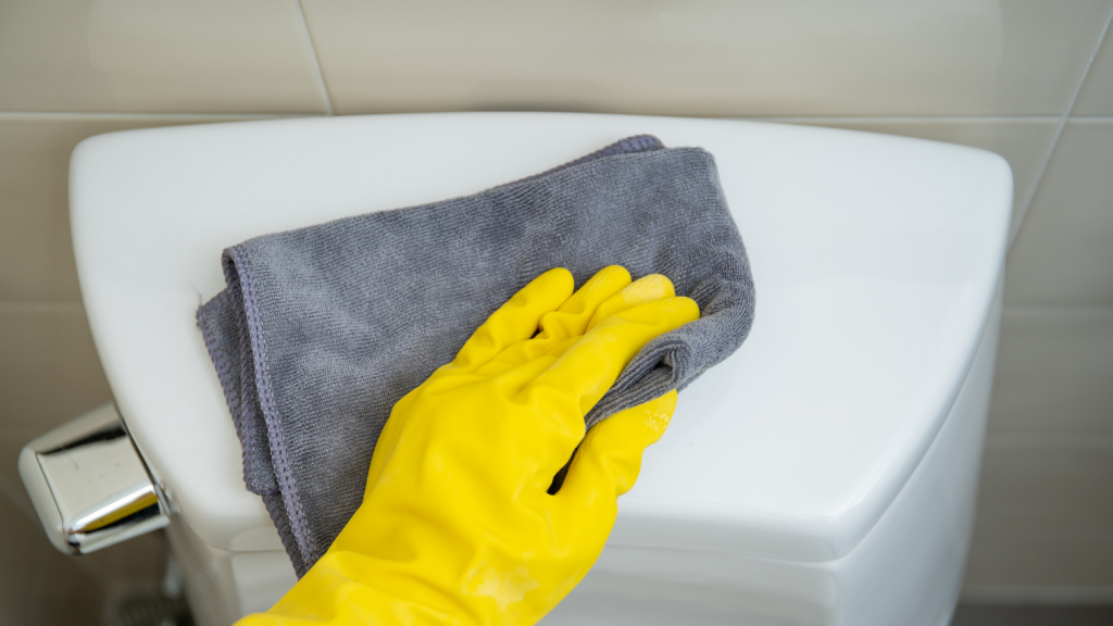 Hand in a rubber yellow glove wiping a white toilet bowl with a grey microfibre cloth