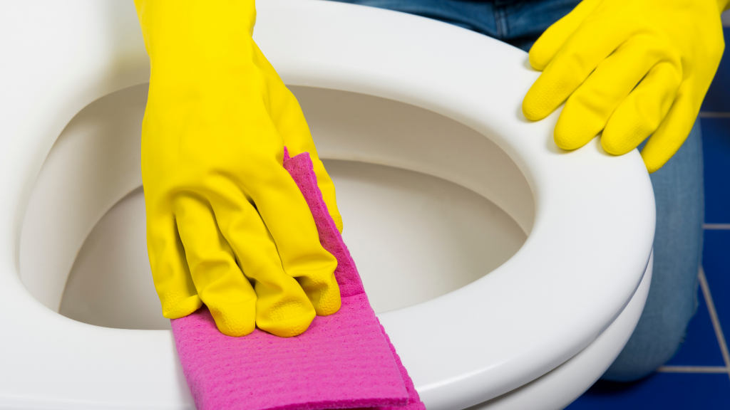 Hands in yellow rubber gloves using a fuchsia cloth to wipe a white toilet seat