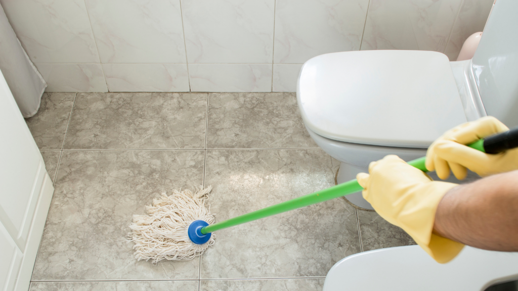 Hands in yellow gloves using a green mop on toilet tiles