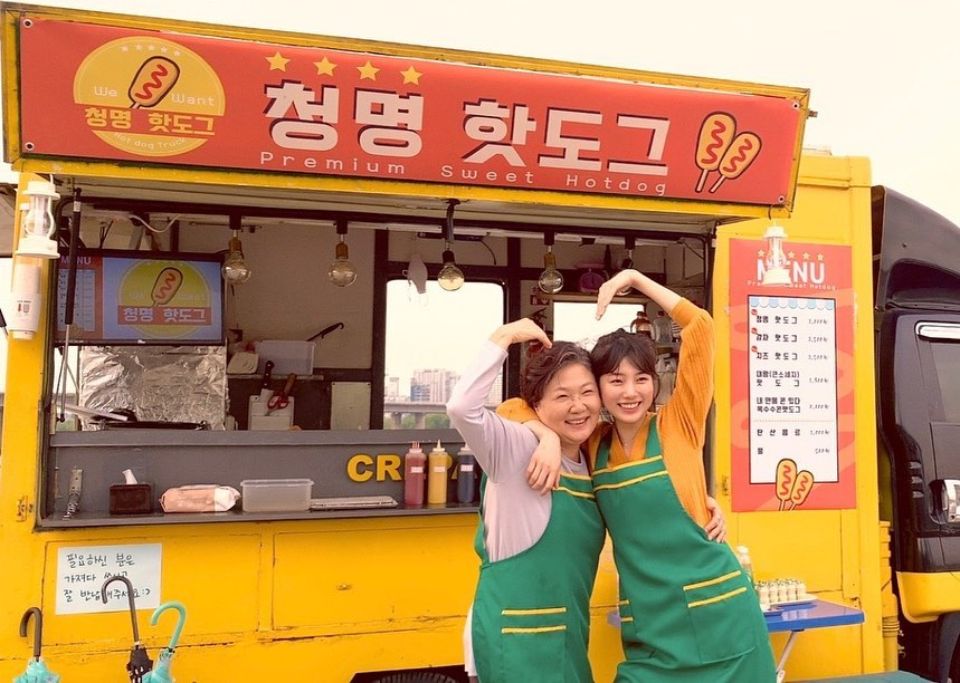 Two Asians standing in front of a yellow corn dog truck with a red Korean signboard