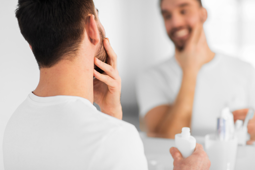 Man holding his beard with one hand, shaving cream in another hand looking at a mirror with a smile